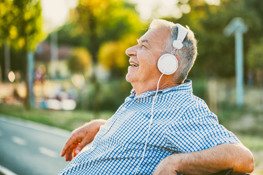 A senior man sitting on a park bench with headphones on.