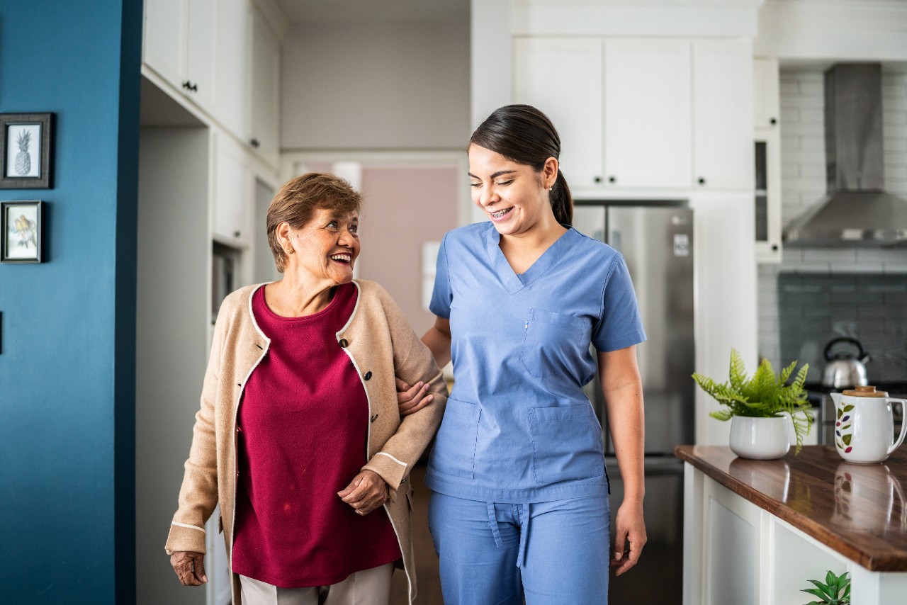 A health care worker holds the arm of an older woman while she walks through her home