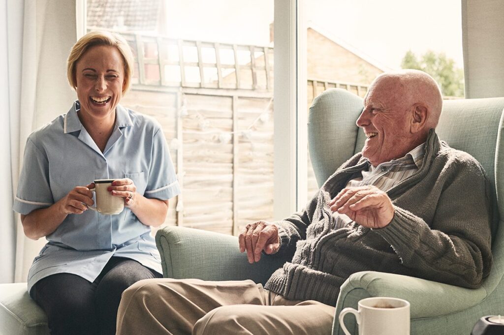 A senior man and nurse laugh over coffee.